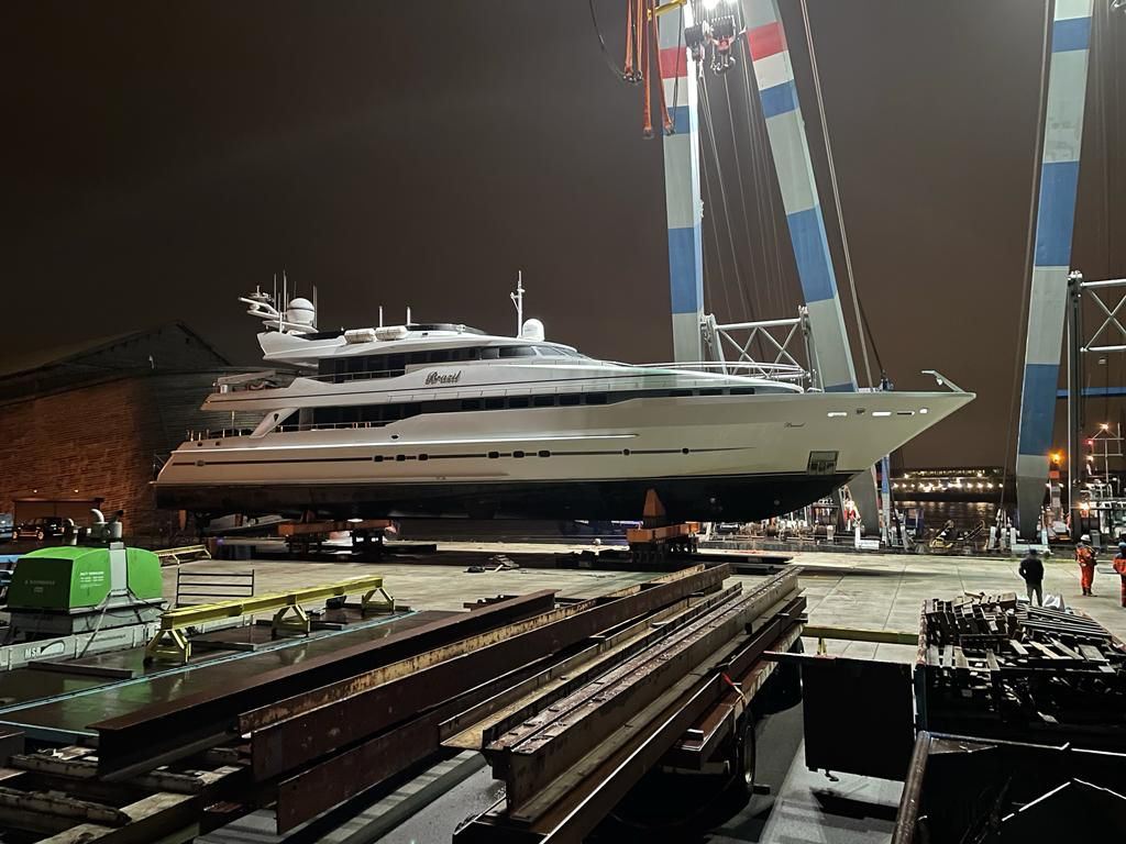 A large motor yacht on transporters at night beside a heavy-lift crane